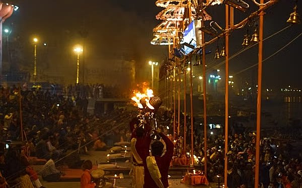 Evening Ganga aarti at Dashashwamedh Ghat: a priest raising a flame lamp beneath the lit umbrella canopies, with the crowd banked up the steps